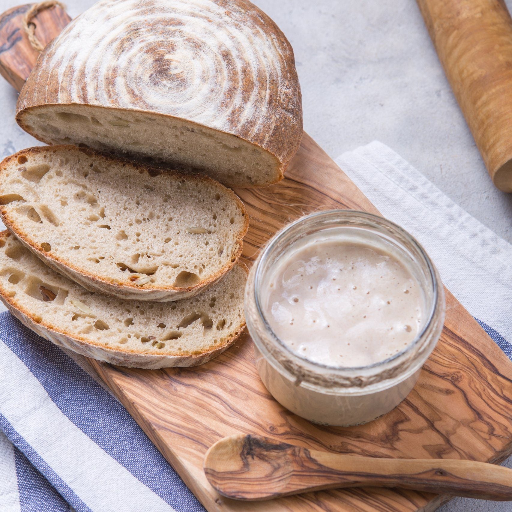 Sliced sourdough loaf beside a jar of bubbly starter on an olive wood board