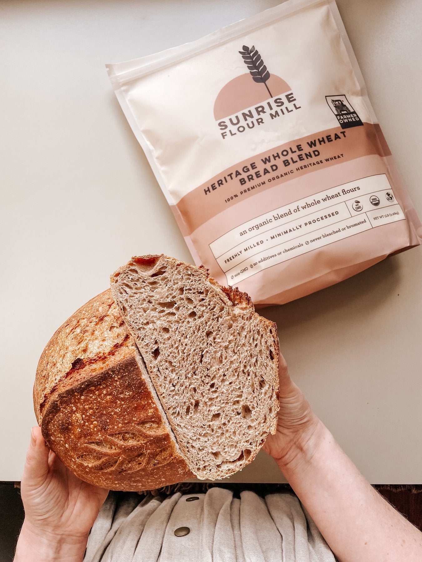 Hands holding a halved sourdough loaf beside a bag of Sunrise flour